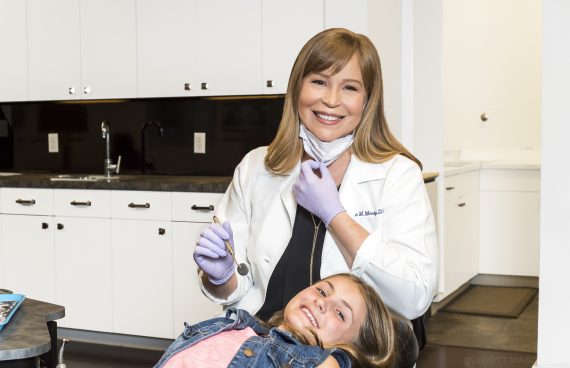 a portrait of a dentist smiling, with a patient in the chair next to her