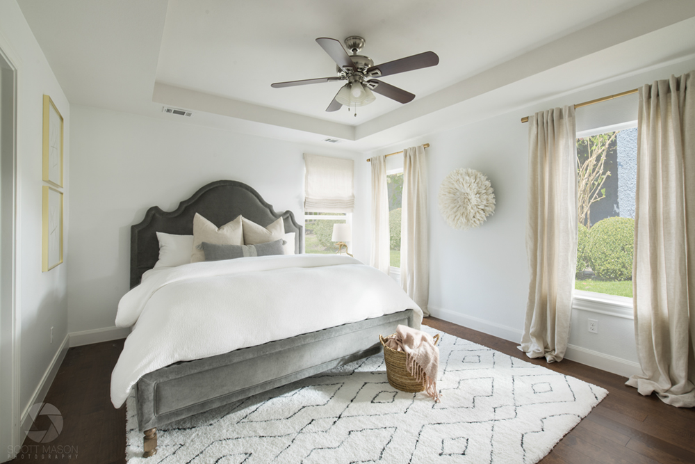 an interior photograph of a white room with a carpet and bed, and a view of shrubs outside