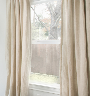 a crop of a window with beige curtains and a fence and tree outside