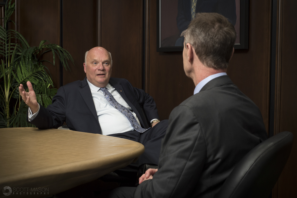 two men in suits sitting at a desk, chatting