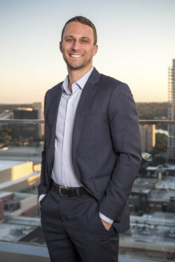 a business portrait/headshot of a smiling man standing on a rooftop downtown